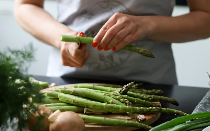 Person with red nail polish peeling green asparagus stalks on a wooden chopping board, with onions and green vegetables nearby, preparing food in a kitchen setting. Keywords: used cars, new cars.