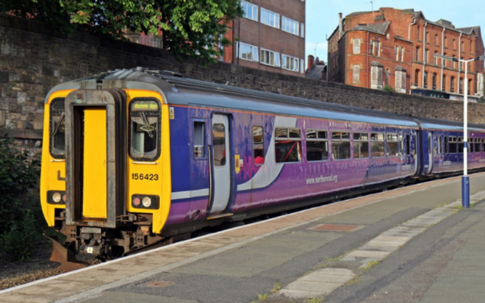 A Northern Rail passenger train with the number 156423 on the front, stopped at a platform with a stone wall and brick buildings in the background. The train is painted purple with yellow front ends and has 'www.northernrail.org' written on its side. The image shows no used cars or new cars, focusing instead on the train and railway station.