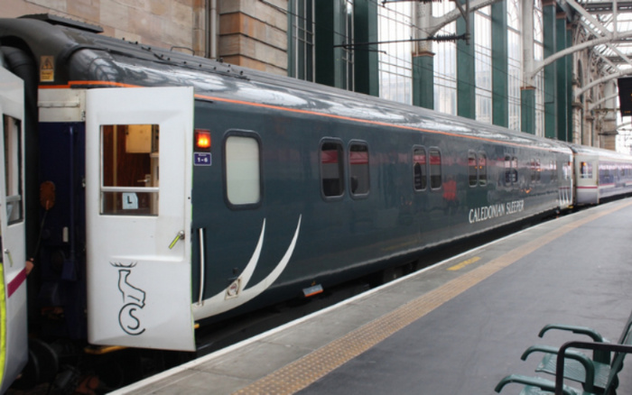 Caledonian Sleeper train at a station platform with open carriage door showing the Caledonian Sleeper logo featuring a stag. The train is dark grey with white branding and is under a large glass and steel station roof, with benches on the platform. No references to used cars or new cars visible in the image.