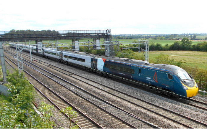 Avanti West Coast high-speed train travelling on railway tracks in a rural area with greenery and fields in the background, with an overhead bridge and electric lines visible, no used cars or new cars present in the image.