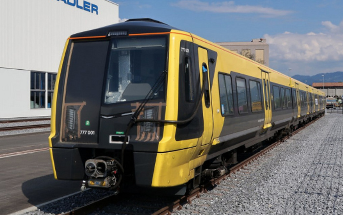 Yellow and black modern train numbered 777 001 parked on railway tracks outside a building with the partial word 'ADLER' visible. The train is stationary on a clear day with mountains in the background, unrelated to used cars or new cars.
