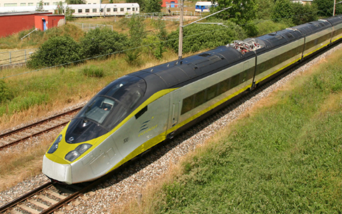 Eurostar high-speed train traveling on railway tracks surrounded by greenery and bushes, with fields and a red building in the background. The train has a sleek silver and yellow design with the number 5001 and the Eurostar logo on its side. Keywords used cars, new cars.