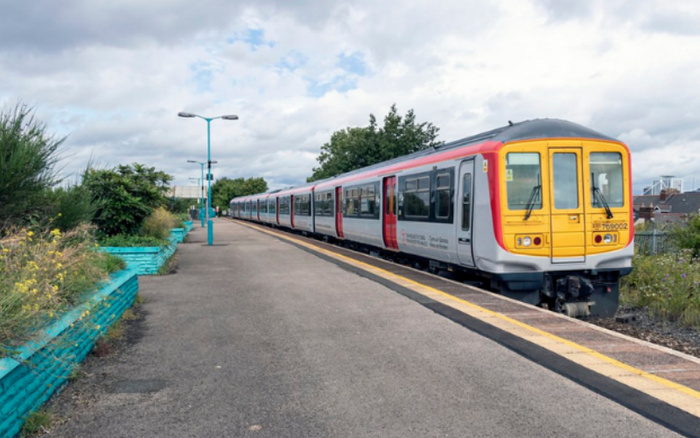 A Northern train at a quiet outdoor railway platform under a cloudy sky, featuring a yellow front and silver body with red accents. The station is empty, surrounded by greenery and blue-painted retaining walls. No used cars or new cars are visible in the image.