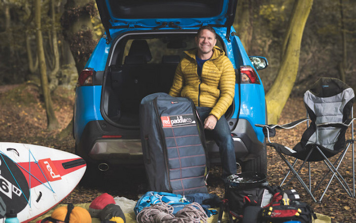 Smiling man in a yellow jacket sitting at the open boot of a blue car in a wooded area, surrounded by outdoor gear including a paddleboard labeled 'red paddle co', ropes, a folding chair, and various bags, depicting a scene of outdoor adventure and emphasising the suitability of used cars and new cars for active lifestyles.