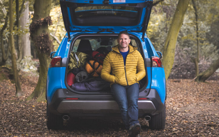 Man in a yellow jacket sitting at the open boot of a blue SUV filled with camping gear in a forest setting, illustrating practical use of cars for outdoor activities, relevant for used cars and new cars buyers interested in versatility and cargo space.