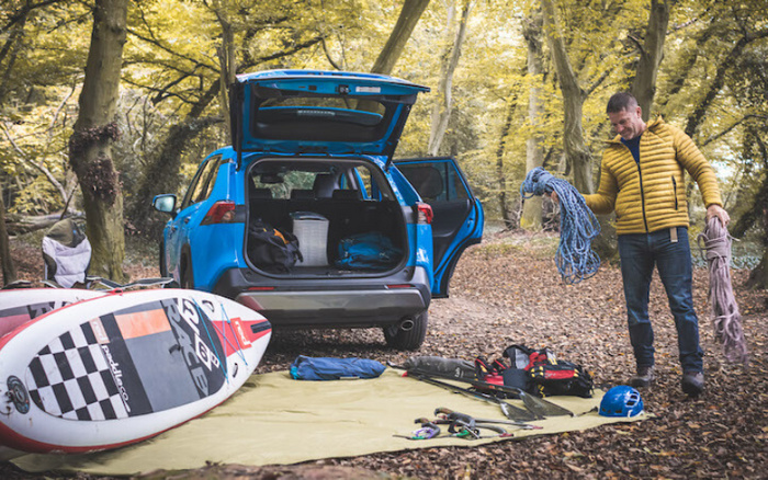 Blue SUV with rear hatch and side door open parked in a forest setting, surrounded by outdoor and climbing gear including a paddleboard, ropes, helmet, and life jackets spread out on a yellow groundsheet, man in a yellow jacket organising climbing ropes nearby, illustrating the versatility of used cars and new cars for outdoor adventures.