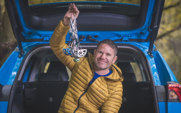 Man in a yellow jacket sitting in the open boot of a blue car, holding up several climbing ascenders. The car is parked outdoors with trees visible in the background, representing an active lifestyle associated with used cars or new cars for recreational purposes.