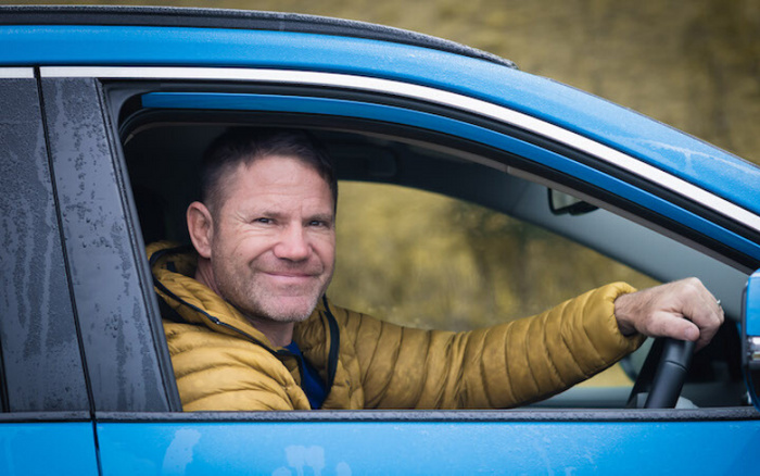 Man in yellow jacket smiling while sitting in the driver's seat of a blue car with the window down on a rainy day, representing used cars or new cars.