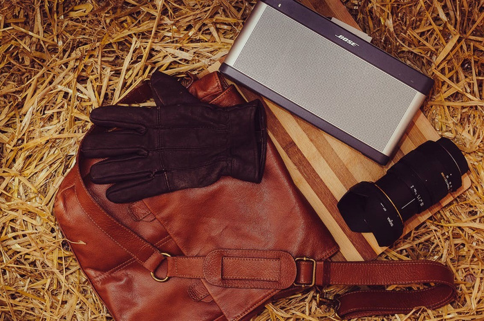 Brown leather bag, black leather gloves, Bose portable speaker, and a Sigma camera lens placed on a wooden board, all set on a bed of straw, captured in a flat lay style photo.