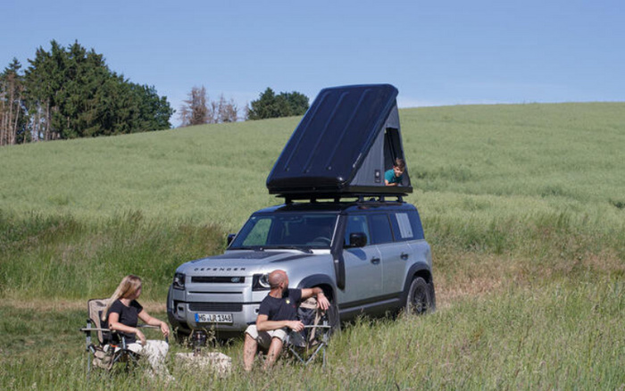 Silver Land Rover Defender parked in a grassy field with its roof tent open. A man and woman sit in camping chairs next to the vehicle, enjoying the outdoors, while a child looks out from the elevated roof tent. The scene depicts an outdoor adventure with a new car suitable for camping and exploration, highlighting both new cars and used cars for outdoor lifestyles.