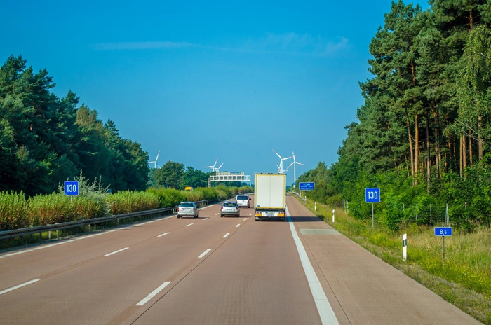 Highway scene with several vehicles including cars and a truck driving on a three-lane road surrounded by greenery and trees under a clear blue sky. Road signs display '130' indicating the speed limit and '8.5' marking a distance. Wind turbines and a distant building are visible ahead, representing a modern, clean environment suitable for new cars and used cars travel and transport.