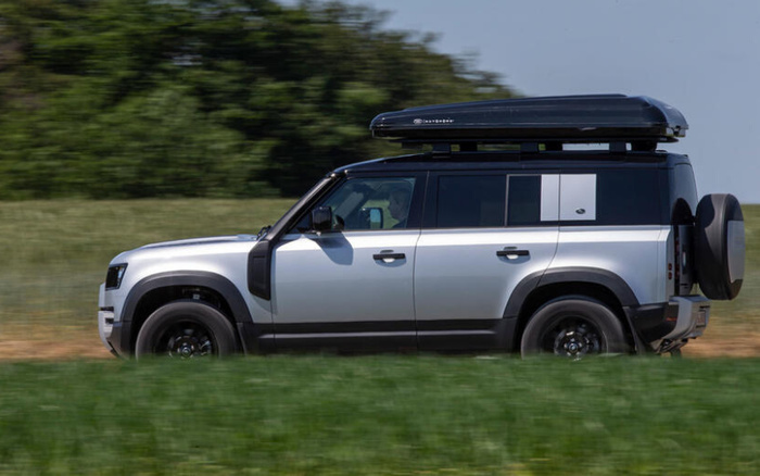 Silver and black Land Rover Defender driving on a grassy path with a black roof storage box labelled 'WAGTOP' attached, set against a background of trees and blue sky, representing used cars and new cars in off-road adventure style.