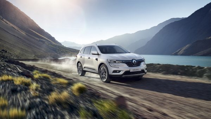 Silver Renault Koleos SUV driving on a gravel road alongside a lake surrounded by mountains on a clear sunny day, representing new cars suitable for rugged terrain, ideal for buyers interested in used cars and new cars.