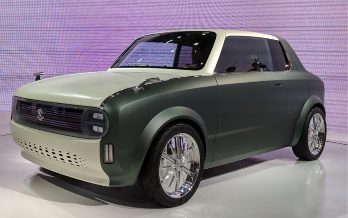 Green and white Suzuki compact car displayed indoors on a white platform with a purple pixelated backdrop. The car has a retro-modern design with smooth, rounded edges, large chrome wheels, and a distinctive front grille featuring the Suzuki logo. This vehicle represents a new car model, suitable for marketing used cars or new cars.