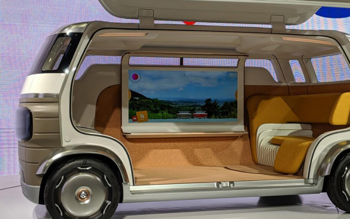 Interior view of a modern, futuristic vehicle with an open side showing a spacious seating area featuring mustard yellow and white seats. The vehicle has a large screen displaying an outdoor scene with two vans and a sky backdrop. The design showcases innovative automotive concepts relevant to new cars and could appeal to buyers of used cars interested in the latest models.