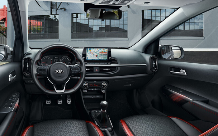 Interior view of a Kia car showcasing the steering wheel with Kia logo, central dashboard featuring a touchscreen displaying a navigation map, air conditioning controls, and manual transmission gear stick. The image highlights modern car features suitable for used cars and new cars buyers. The seats have a geometric pattern with red accents, and the windows reveal an industrial building exterior.