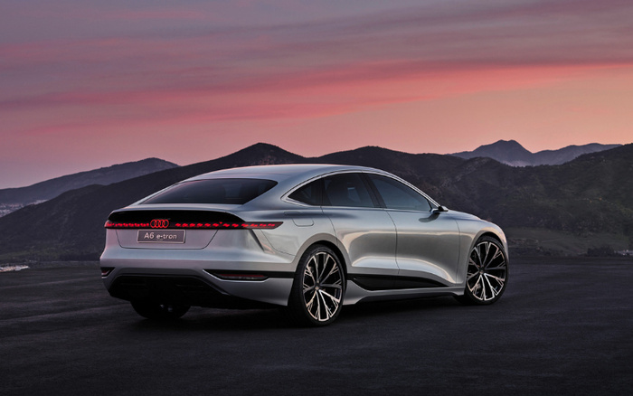 Silver Audi A6 e-tron parked on a dark asphalt surface with mountains in the background under a pink and purple sunset sky, showcasing a sleek design with black tinted windows, large alloy wheels, and a continuous rear LED light strip. Ideal image for used cars and new cars listings.
