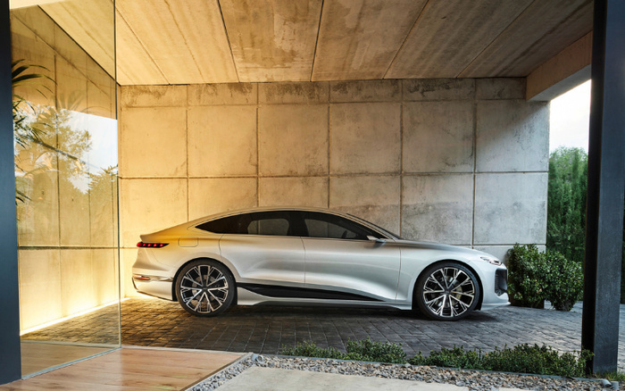 Silver modern electric car parked under a concrete roof with a large glass wall on the left and greenery on the right, showcasing a sleek design, ideal for advertising new cars alongside used cars.