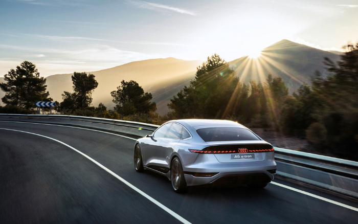 Silver Audi A6 e-tron driving on a curved mountain road during sunset with sunlight filtering through trees and hills, showcasing a modern new car model relevant for used cars and new cars market.