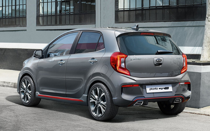 Rear and side view of a grey Kia Picanto GT-Line parked on a street in front of a building with large windows. The compact hatchback features stylish alloy wheels, distinctive rear lights, and a sporty design with red accents along the lower body panels and rear bumper. The image showcases the car's modern appearance, suitable for advertising used cars and new cars for sale.