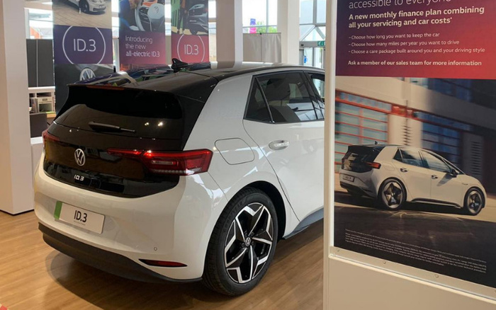 White Volkswagen ID.3 electric car displayed indoors at a dealership showroom, with promotional banners in the background featuring the text 'Introducing the new all-electric ID.3' and a large poster highlighting a monthly finance plan combining servicing and car costs, emphasizing flexible terms for keeping the car, mileage limits, and care package options. The scene represents both new cars and used cars financing solutions.