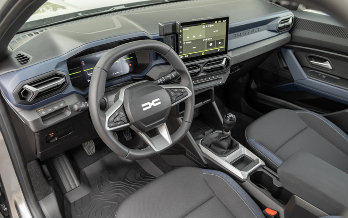 Interior view of a Dacia Duster new car showcasing the driver's seat, steering wheel with Dacia logo, digital dashboard, and central touchscreen displaying media controls. The car features a manual gear stick, modern air vents, and a clean, modern design, ideal for promotion in used cars and new cars market.