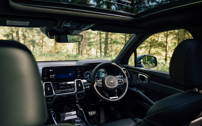 Interior view of a modern Kia car featuring a digital dashboard displaying the time 2:14 PM and weather as 6°C, a multi-function steering wheel with the Kia logo, black leather seats, a panoramic sunroof with a partial open view, and a sleek centre console with touch controls. The scene outside shows a forest setting, highlighting the contrast between the high-tech interior and nature. This image is optimised for used cars and new cars dealerships highlighting advanced car interiors.