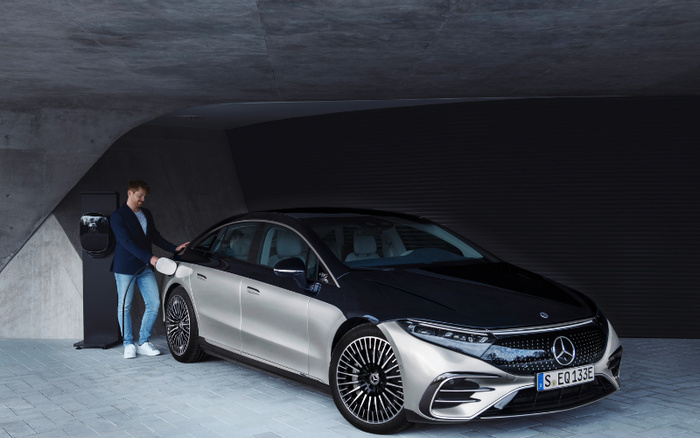 A man in a dark blazer and jeans charging a silver and black Mercedes-Benz EQS electric car at a modern charging station in a sleek, covered parking area. The car's license plate reads S EQ 133E. The scene implies the availability of used cars and new cars including electric models.