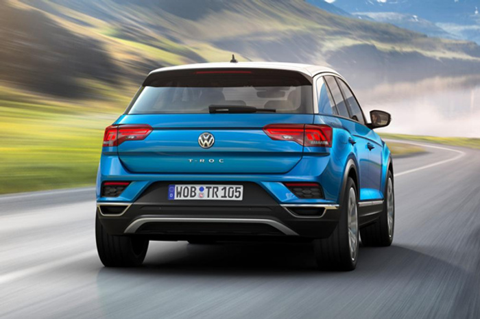 Rear view of a blue Volkswagen T-Roc driving on a winding road through a mountainous landscape under a partly cloudy sky. The vehicle displays a European license plate with the characters WOB TR 105. Ideal for used cars and new cars buyers seeking a stylish compact SUV.