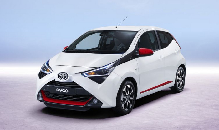 White Toyota Aygo compact car with red accents on the front grille, side mirrors, and side trim, displayed in a studio setting with a light purple floor and a light blue background, ideal for advertising used cars and new cars.