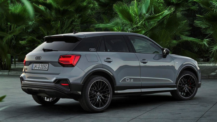 Grey Audi Q2 parked on a tiled surface with lush green palm trees in the background. The car has black alloy wheels, tinted windows, and the license plate reads 'IN-Q 2050'. Suitable for illustrating used cars and new cars.