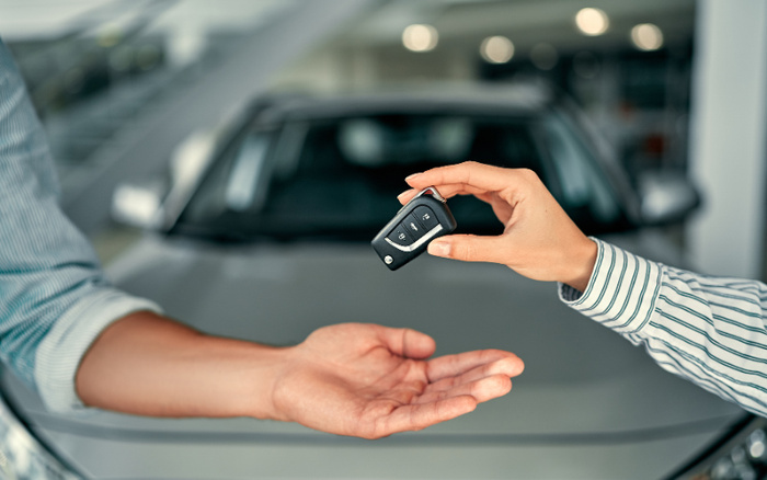 A close-up of two people exchanging a car key in front of a car, symbolising the sale or handover of a vehicle. One hand is holding a black car key with buttons, ready to pass it to the other open hand. The blurred car in the background suggests a dealership or showroom setting, relevant to buying used cars or new cars.