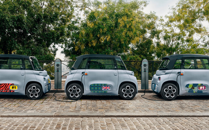 Three compact electric cars parked side by side and connected to charging stations on a cobbled street with green trees in the background. Each car is light grey with black roofs and features unique colourful decal designs on their doors and rear panels, including a zebra pattern with a #1 badge, a British flag on a blue plaid pattern, and a camouflage pattern with a #1 badge. The scene highlights eco-friendly used cars and new cars integrating electric vehicle technology.