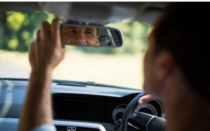 Man adjusting the rearview mirror inside a car, focusing on his reflection; interior details include the steering wheel and dashboard, representing the experience of driving new cars or used cars.