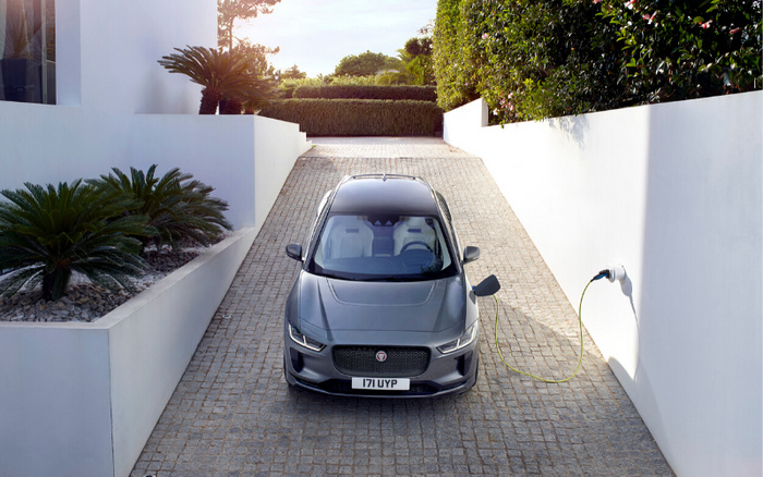 Front view of a grey Jaguar electric car parked on a cobblestone driveway, connected to a white electric vehicle charger fixed on a white wall. Green plants and palm trees line the side of the driveway leading to a modern house. The car's number plate reads 171 UYP. Ideal image for used cars and new cars in the electric vehicle category.