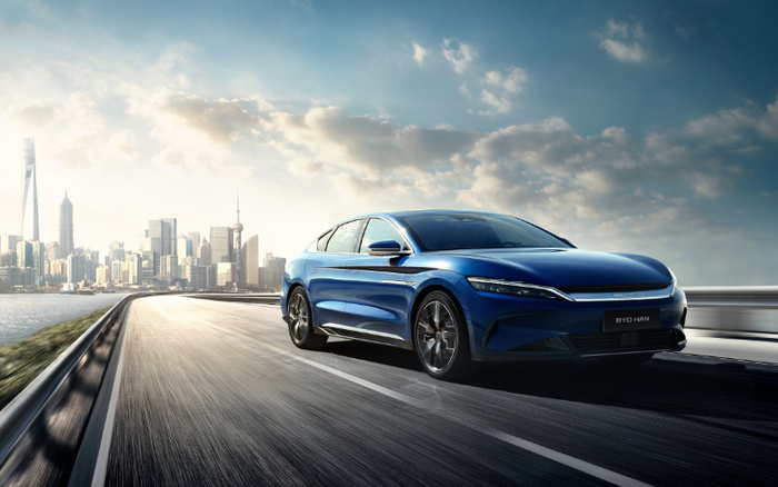 Blue BYD Han electric car driving on a highway with a modern city skyline in the background under a partly cloudy sky, showcasing sleek design suitable for those interested in new cars and used cars.