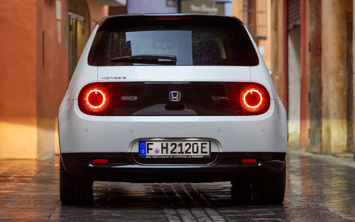 Rear view of a white Honda e electric car with distinctive round red tail lights illuminated, parked on a wet cobblestone street between orange and yellow buildings. The car has a German license plate reading 'F H 2120E' and a small text below the plate says 'Gute Fahrt wünscht Ihr Autohaus am Frankfurter'.