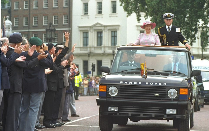 A black Range Rover driving down a city street with Queen Elizabeth II standing through the sunroof wearing a pink dress and hat, accompanied by a man in a naval uniform. A crowd of veterans and onlookers are clapping and waving from the side of the road. The scene is set in an urban area with white multi-storey buildings in the background. The image represents a classic Range Rover model, relevant to used cars and new cars in terms of luxury and vintage vehicle markets.