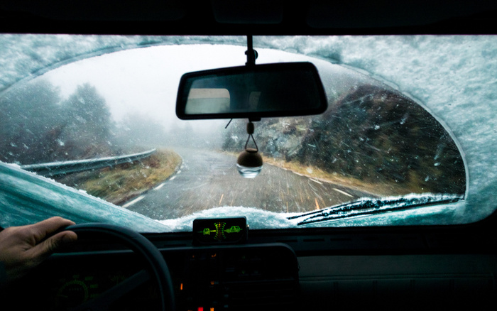 View from inside a car driving on a snowy, winding road with the windshield partially cleared of snow by the wiper. A hand is on the steering wheel and an air freshener hangs from the rearview mirror. The scene highlights the importance of safe driving conditions for used cars and new cars in winter weather.