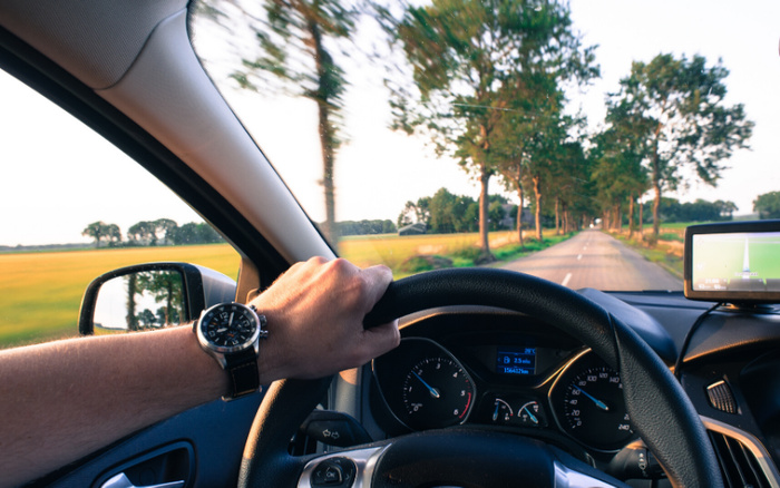 View from the driver's seat of a car on a countryside road lined with trees. The driver's left hand, wearing a black wristwatch, is on the steering wheel. The car dashboard shows a speedometer, fuel gauge, and a digital display with mileage of 156412 km. A GPS navigation device is mounted on the right side of the dashboard. Ideal image for used cars and new cars driving experience and vehicle interior features.