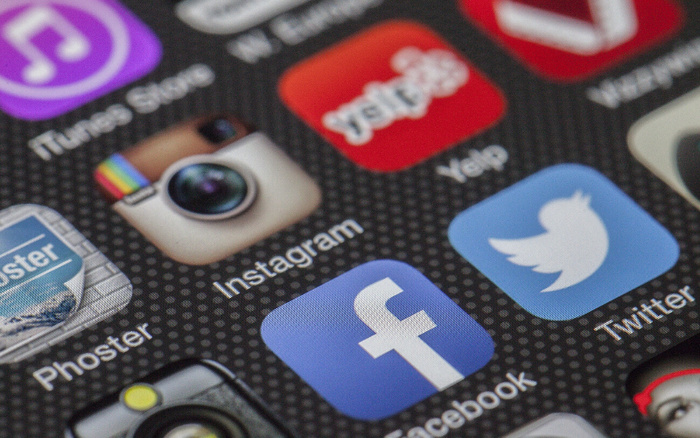 Close-up image of a smartphone screen displaying app icons including iTunes Music, Instagram, Yelp, Phoster, Facebook, and Twitter on a dark textured background, highlighting popular social media and service apps.