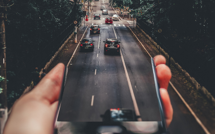 A hand holding a smartphone displaying a live view of a busy urban road with several cars driving, blending seamlessly with the actual street scene. The image highlights the contrast and connectivity between digital technology and real-world driving, relevant to used cars and new cars.