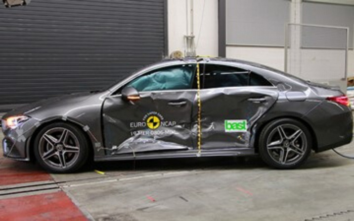 Grey sedan car with significant side damage from crash testing, featuring Euro NCAP and Best logos on the doors, parked inside a testing facility with a black roller shutter door in the background. The car has deployed airbags visible through the windows and is used in safety and crash test assessments for both used cars and new cars.