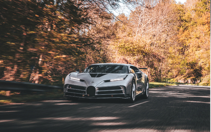 Silver Bugatti sports car driving on a winding forest road during autumn with trees showing fall colours, representing new cars and used cars in a scenic outdoor setting.