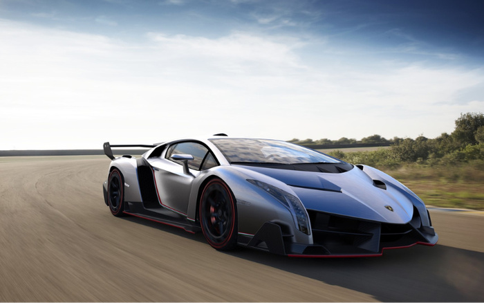 Silver Lamborghini Veneno sports car driving on a curved race track under a clear blue sky with some wispy clouds, surrounded by greenery. The car features sharp aerodynamic design, black and red detailing on the rims and body, and a large rear wing. Ideal image representing high-performance new cars or luxury used cars.