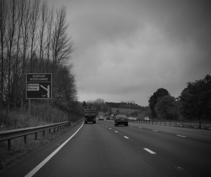 Black and white image of a motorway with several vehicles including cars and a truck traveling on it. Road sign on the left indicates 'Burpham Interchange' with directions to Burpham and Merrow, ½ mile ahead. Surrounding scenery shows leafless trees and some foliage under a cloudy sky. Keywords focused on used cars and new cars on the road.