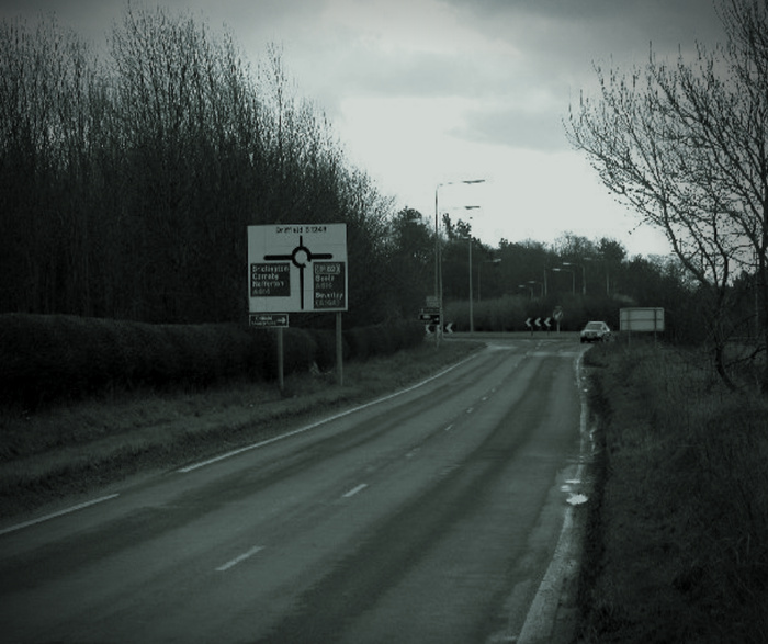 Road leading to a roundabout with a directional sign indicating destinations including Bicester, Cirencester, Moreton-in-Marsh, Aston, Banbury, and Drayton. The sign is positioned alongside a quiet rural road bordered by trees and bushes, with a car visible approaching the roundabout. Scene captures a typical British countryside road, relevant for used cars and new cars travel routes.