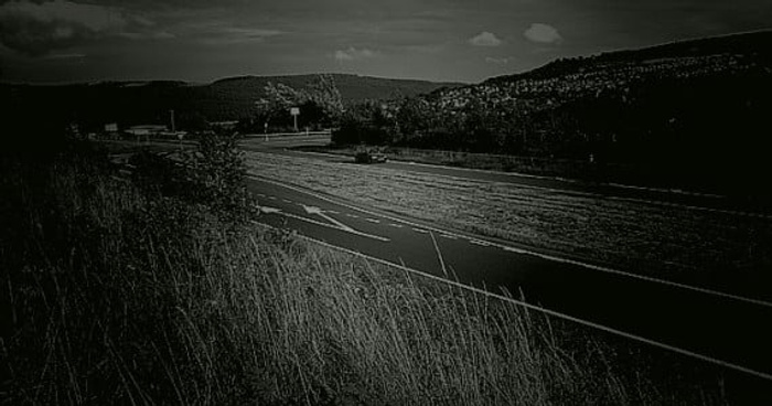 Roadside view with dry grass in the foreground and hills in the background under a partly cloudy sky. No visible cars or text in the image.