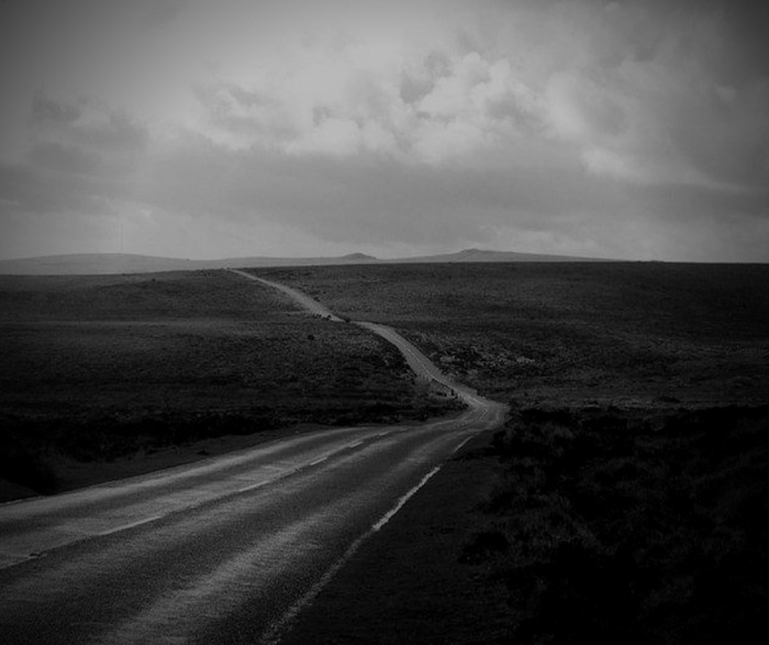Black and white image of a long, empty road stretching into a barren landscape under a cloudy sky, with no visible text or references to used cars or new cars.