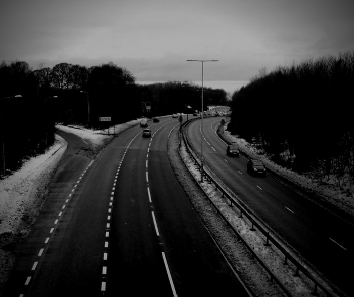 Black and white image of a divided highway with several cars driving on it, surrounded by trees and light snow on the ground, no visible text, depicting a typical road scene suitable for illustrating used cars and new cars in transit.